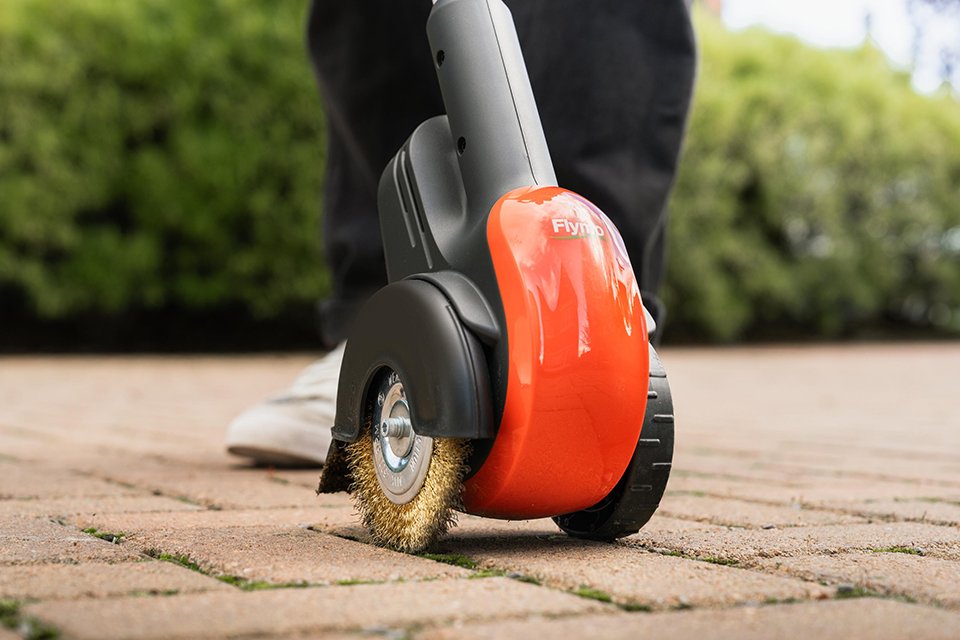 A person using a Flymo® 18V EasiWeeder weed brush to remove garden weeds.